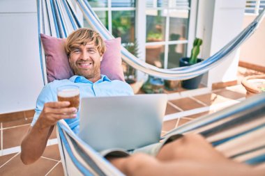 Evinin terasındaki orta yaşlı yakışıklı adam hamakta uzanıp dizüstü bilgisayarla çalışıyor ve taze bira içiyor.