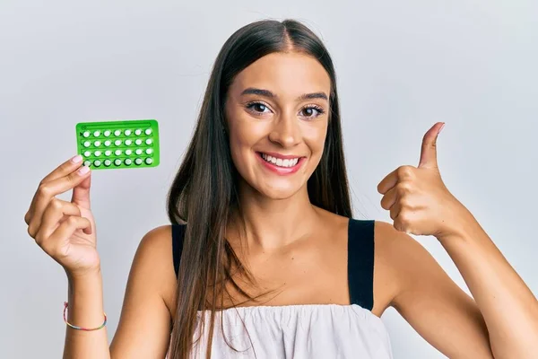 Young hispanic woman holding birth control pills smiling happy and positive, thumb up doing excellent and approval sign