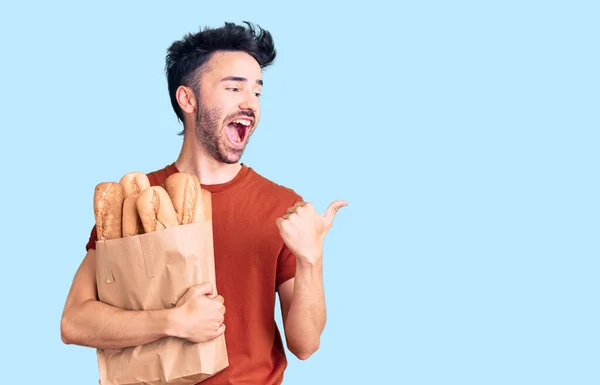 Young Hispanic Man Holding Paper Bag Bread Pointing Thumb Side — Stock Photo, Image