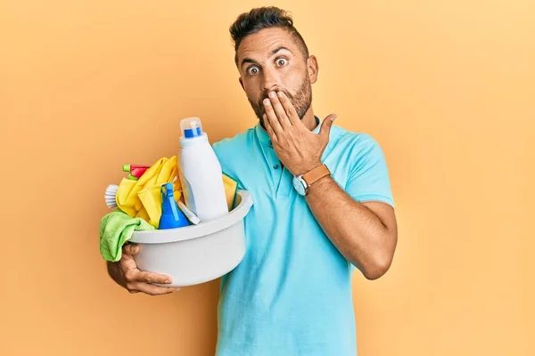 Handsome man with beard holding cleaning products covering mouth with hand, shocked and afraid for mistake. surprised expression