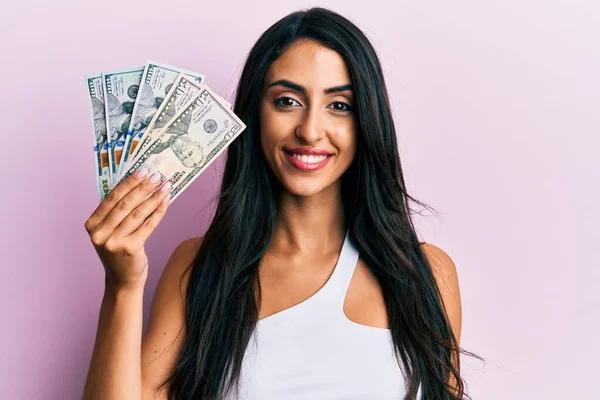 Beautiful hispanic woman holding dollars looking positive and happy standing and smiling with a confident smile showing teeth