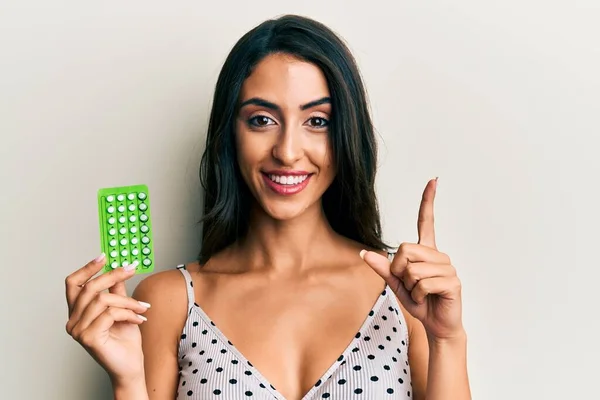 Beautiful hispanic woman holding birth control pills smiling with an idea or question pointing finger with happy face, number one