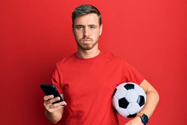 Handsome caucasian man holding football ball looking at smartphone puffing cheeks with funny face. mouth inflated with air, catching air. 