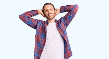 Rahat ve esnek giyinen genç yakışıklı adam, kolları ve elleri başının arkasında mutlu bir şekilde gülümsüyor. 