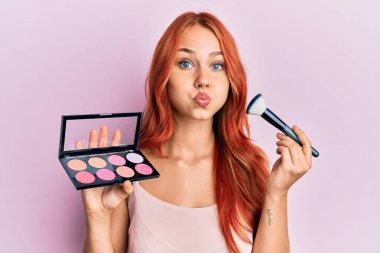 Young redhead woman holding makeup and brush puffing cheeks with funny face. mouth inflated with air, catching air.  clipart