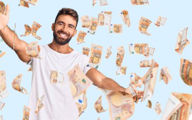 Latin asıllı genç bir adam, günlük beyaz tişört giyip kameraya bakıyor ve sarılmak için kollarını açmış gülümsüyor. Mutluluğu kucaklayan neşeli bir ifade..