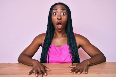Saçları günlük kıyafetlerle örülü Afrika kökenli Amerikalı bir kadın masada korkmuş ve ağzı açık bir şekilde şaşkınlıkla oturuyor. 