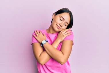 Kısa saçlı, pembe tişörtlü güzel bir kadın pembe arka planda kendine sarılıyor mutlu ve pozitif, kendinden emin gülümsüyor. Sevgi ve özgüven. 