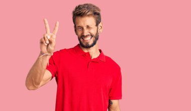 Sakallı, sıradan kıyafetler giyen, kameraya gülümseyen ve zafer işareti yapan yakışıklı sarışın adam. İki numara.. 