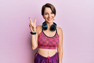 Kısa saçlı, spor giyimli, kulaklık takan genç esmer kadın iki numarayı gösterip işaret ederken kendinden emin ve mutlu gülümsüyor.. 