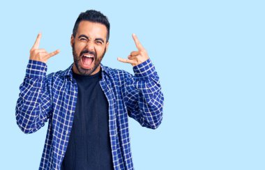 Latin asıllı genç adam, gündelik kıyafetler giyip çılgın bir ifadeyle bağırıyor ve elleri havada rock sembolü yapıyor. Müzik yıldızı. ağır müzik konsepti. 