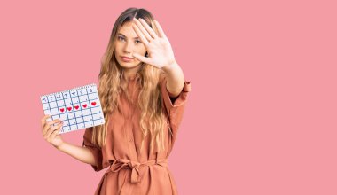 Güzel beyaz kadın, sarı saçlı, elinde kalp takvimiyle dur işareti yapıyor ciddi ve kendinden emin bir ifadeyle, savunma jesti yapıyor. 