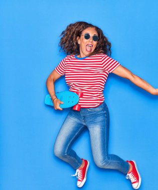 Orta yaşlı, güzel, güneş gözlüğü takan mutlu bir kadın. Yüzünde tebessümle sıçrayıp tenha mavi fırında kaykay yapıyor.