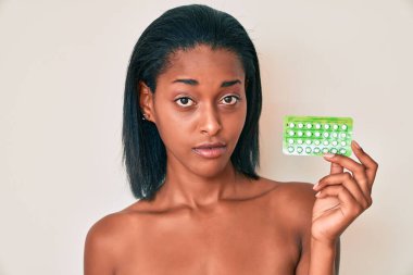 Genç Afro-Amerikalı kadın elinde doğum kontrol hapları tutarken yüzünde ciddi bir ifadeyle rahatladı. Basit ve doğal bir şekilde kameraya bakıyorsun.. 