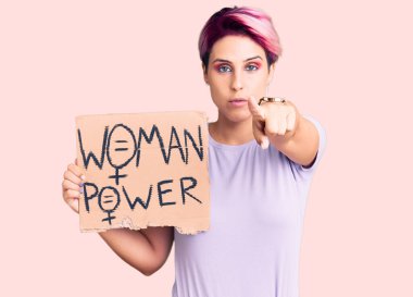Pembe saçlı, elinde kadın pankartı olan genç ve güzel bir kadın kamerayı işaret ediyor. Kendine güvenen bir jest, ciddi görünüyor. 