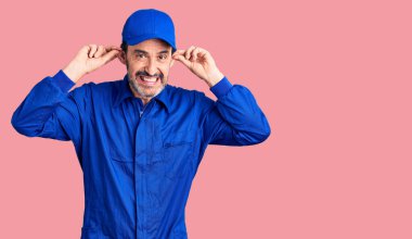 Orta yaşlı yakışıklı, tamirci üniforması giyen, gülümseyen kulakları parmaklarıyla çeken, komik bir jest. deneme sorunu 