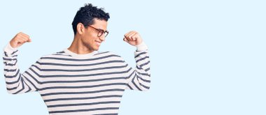 Sıradan kıyafetler ve gözlük takan İspanyol yakışıklı genç adam gururla gülümseyen kol kaslarını gösteriyor. fitness konsepti. 