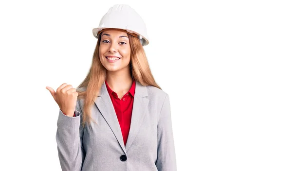 漂亮的年轻女子戴着建筑师的硬礼帽 笑容满面 面带微笑 用大拇指指着旁边 — 图库照片
