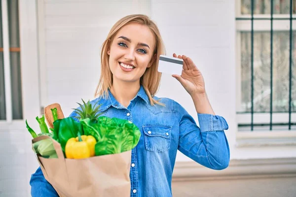 年轻的金发姑娘高兴地微笑着 手里拿着食品杂货纸袋和信用卡站在城里 — 图库照片