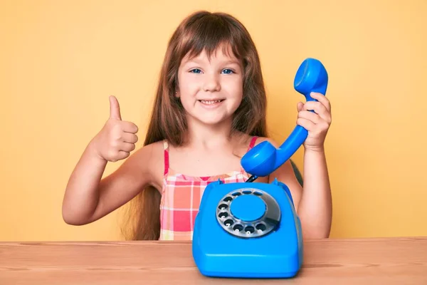 一个有着长发的高加索小女孩坐在桌子上 用老式电话开心而积极地微笑着 竖起大拇指做着出色的事 签了字 — 图库照片