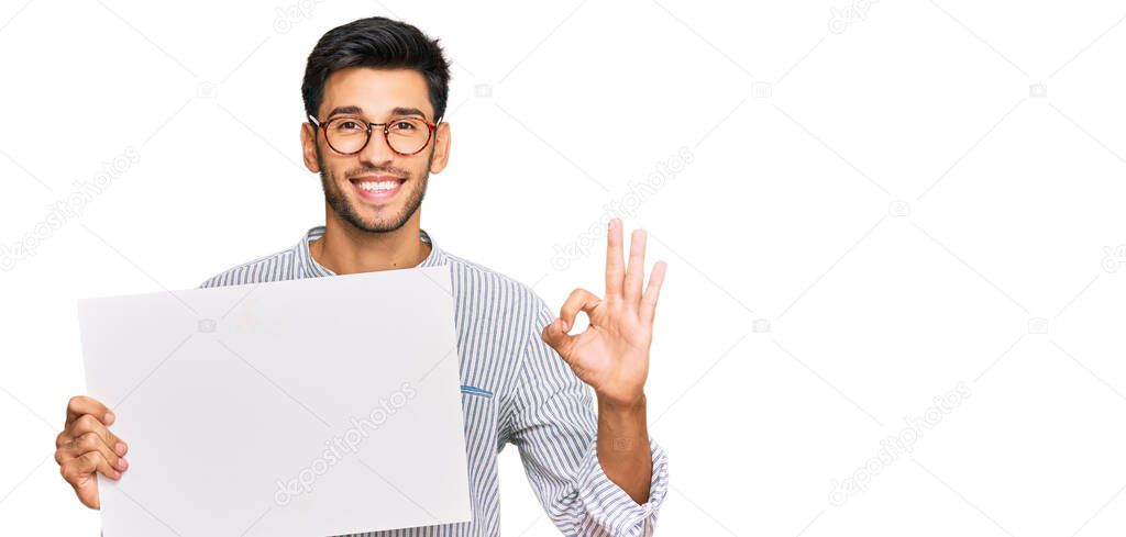 Young handsome man holding blank empty banner doing ok sign with fingers, smiling friendly gesturing excellent symbol 