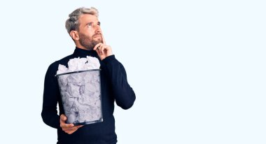 Kırışmış kağıtlarla kağıttan çöp tenekesi tutan genç, yakışıklı sarışın adam ciddi bir yüz ifadesiyle eli çenesinde soru soran, kafa karıştırıcı bir fikir hakkında düşünceli. 