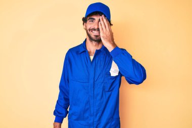 Kıvırcık saçlı ve ayı gibi giyinmiş yakışıklı genç bir adam. Tulum giymiş, bir gözünü elinle kapatıyor. Yüzünde kendine güvenen bir gülümseme ve şaşırtıcı bir duygu.. 