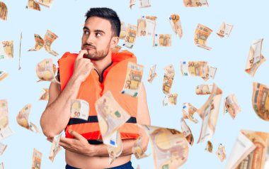 Can yeleği giymiş genç yakışıklı bir adam ciddi bir yüz ifadesi, eli çenesinde soru soran, kafa karıştırıcı bir fikir hakkında düşünceli.