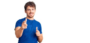 Kıvırcık saçlı, sıradan giyinen, mutlu ve komik yüzlü genç yakışıklı adam kamerayı işaret ediyor. iyi enerji ve titreşimler. 