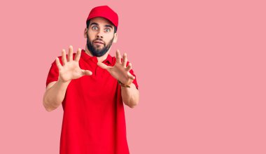 Sakallı, teslimat üniforması giyen, korku ifadesi karşısında dehşete düşmüş genç yakışıklı adam. El hareketini bırak, şok içinde bağır. panik konsepti. 