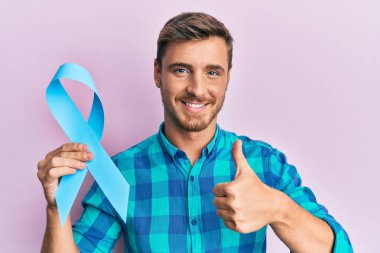 Mavi kurdeleli yakışıklı beyaz adam mutlu ve pozitif gülümsüyor, başparmağıyla mükemmel ve onay işareti yapıyor. 