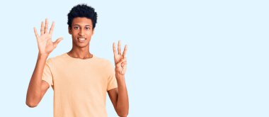 Genç, Afro-Amerikalı, günlük giysiler giymiş, 8 numaralı parmaklarıyla işaret ederken, kendinden emin ve mutlu bir şekilde gülümsüyor.. 