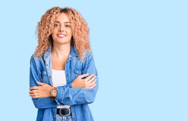 Kıvırcık saçlı, sıradan kot ceket giyen, kollarını kavuşturup kameraya bakan mutlu yüzlü genç sarışın kadın. pozitif kişi. 