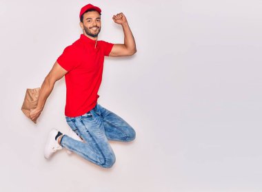 Mutlu gülümseyen üniformalı yakışıklı İspanyol teslimatçı. Yüzünde tebessümle, elinde torbayla, izole edilmiş beyaz arka planda paket yiyeceklerle zıplıyorsun..