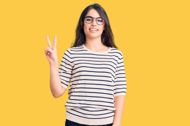 Esmer, sıradan kıyafetler ve gözlük takan genç kız iki numarayı gösterip işaret ederken kendinden emin ve mutlu bir şekilde gülümsüyor.. 