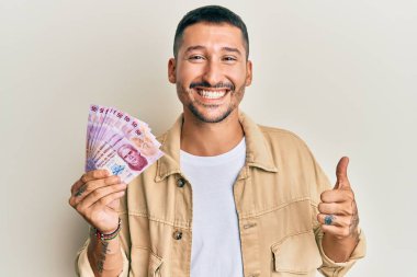 50 Meksika pezosu tutan yakışıklı adam mutlu ve olumlu gülümsüyor, başparmağıyla mükemmel ve onay işareti yapıyor. 