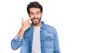 Latin asıllı genç bir adam, gündelik giysiler giyip el ve burnu işaret ediyor, neşeli bir şekilde gülümsüyor. güzellik konsepti 