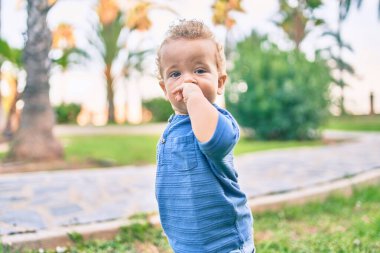 Üzgün küçük çocuk, güneşli bir günde parkta diş etlerine dokunduğu için parmaklarını dudaklarına koyuyor. Güzel sarı saçlı erkek bebek yeni bebek dişleri için acı çekiyor.