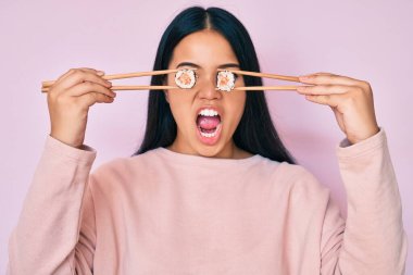 Genç ve güzel Asyalı bir kız yemek çubuklarıyla gözlerine suşi sürüyor kızgın ve öfkeli çığlıklar atıyor ve öfkeyle bağırıyor. öfke ve agresif konsept. 