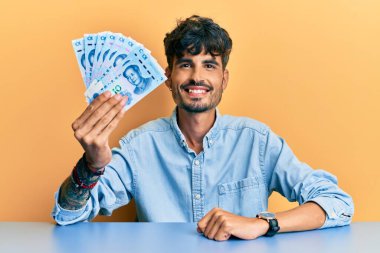 Genç İspanyol adam Yuan Çin banknotlarını tutuyor masaya oturmuş mutlu ve pozitif görünüyor ve gülümsüyor dişlerini göstererek gülümsüyor 