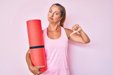 Orta yaşlı İspanyol kadın kızgın suratlı yoga minderi tutuyor, negatif işaret baş parmakları aşağıda, reddedilme konsepti. 