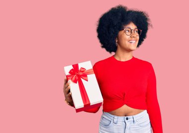 Genç Afro-Amerikalı kız elinde hediyeyle gülümseyerek, doğal bir ifadeyle yan yana bakıyor. Kendinden emin gülen. 