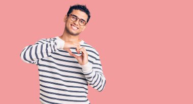 Gündelik giysiler ve gözlük takan, aşık gülümseyen, elleri ile kalp sembolü yapan İspanyol yakışıklı genç adam. romantik kavram. 