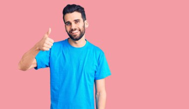 Sakallı, sıradan tişörtlü, mutlu ve pozitif gülümseyen, başparmağıyla mükemmel ve onay işareti yapan genç yakışıklı bir adam. 