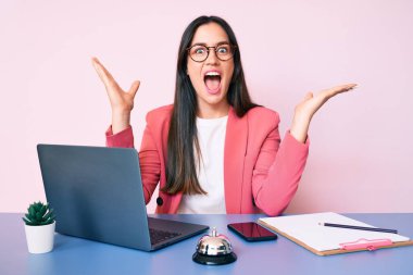 Genç beyaz kadın resepsiyonist masasında oturmuş laptopla zaferi kutluyor gülümseyerek ve ellerini kaldırarak kazanan ifadeyle. 