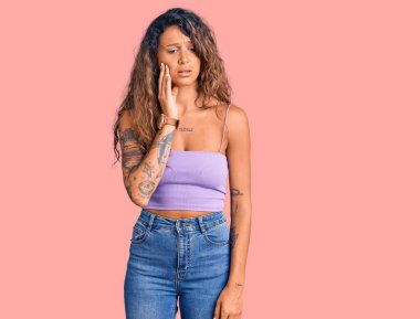 Dövmeli, sıradan kıyafetler giyen, diş ağrısı ya da diş ağrısı yüzünden ağızlarına acı dolu bir ifadeyle dokunan genç İspanyol kadın. dişçi 