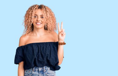 Kıvırcık saçlı, günlük yaz kıyafetleri giyen, kameraya gülümseyen ve zafer işareti yapan genç sarışın kadın. İki numara.. 