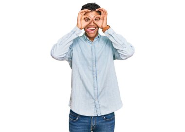 Genç, yakışıklı, Afro-Amerikalı sıradan kıyafetler giyen, dürbün gibi jest yapan, dilini dışarı çıkaran, gözleriyle parmaklarından bakan bir adam. çılgın ifade. 