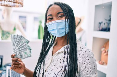 Tıbbi maske takan genç Afro-Amerikalı kadın giyim mağazasında dolar banknotları tutuyor.