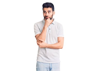 Sakallı, eli çenesinde polo giyen, soru soran, düşünceli, yakışıklı bir genç. Düşünceli bir yüzle gülümsüyor. şüphe kavramı. 